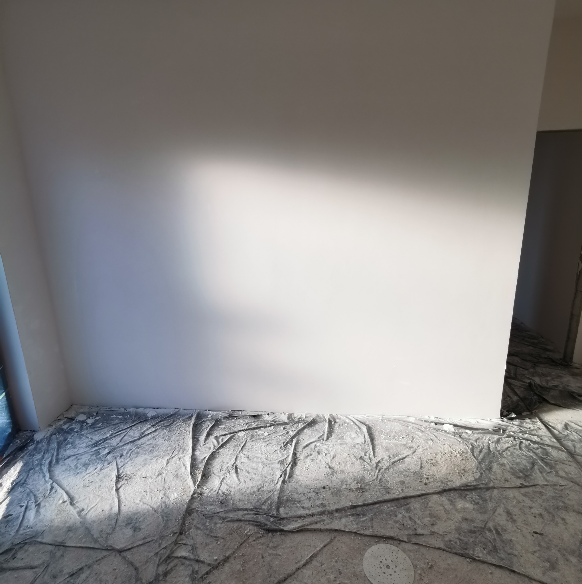 Wnętrze w trakcie prac przygotowawczych: świeżo zagruntowana ściana pod kątem, zabezpieczona folią podłoga z widocznymi resztkami materiałów budowlanych.