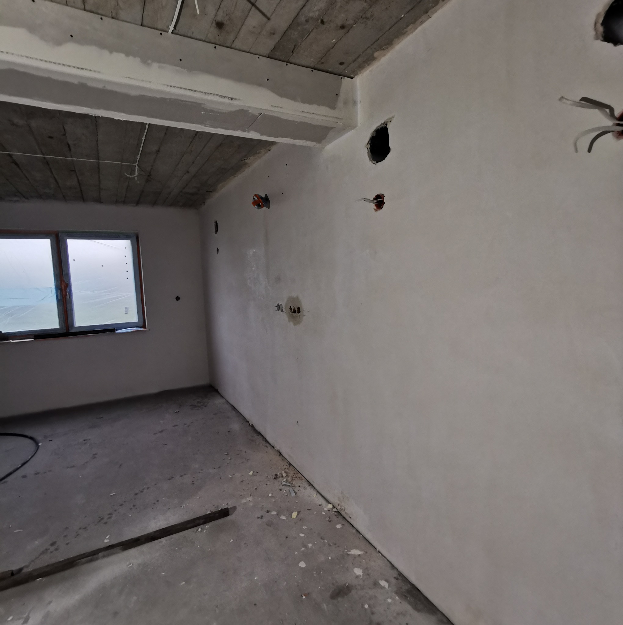 Wnętrze w trakcie prac wykończeniowych: gładkie, białe ściany po nałożeniu, surowy drewniany strop, przygotowane otwory na instalacje elektryczne, okno zabezpieczone folią.