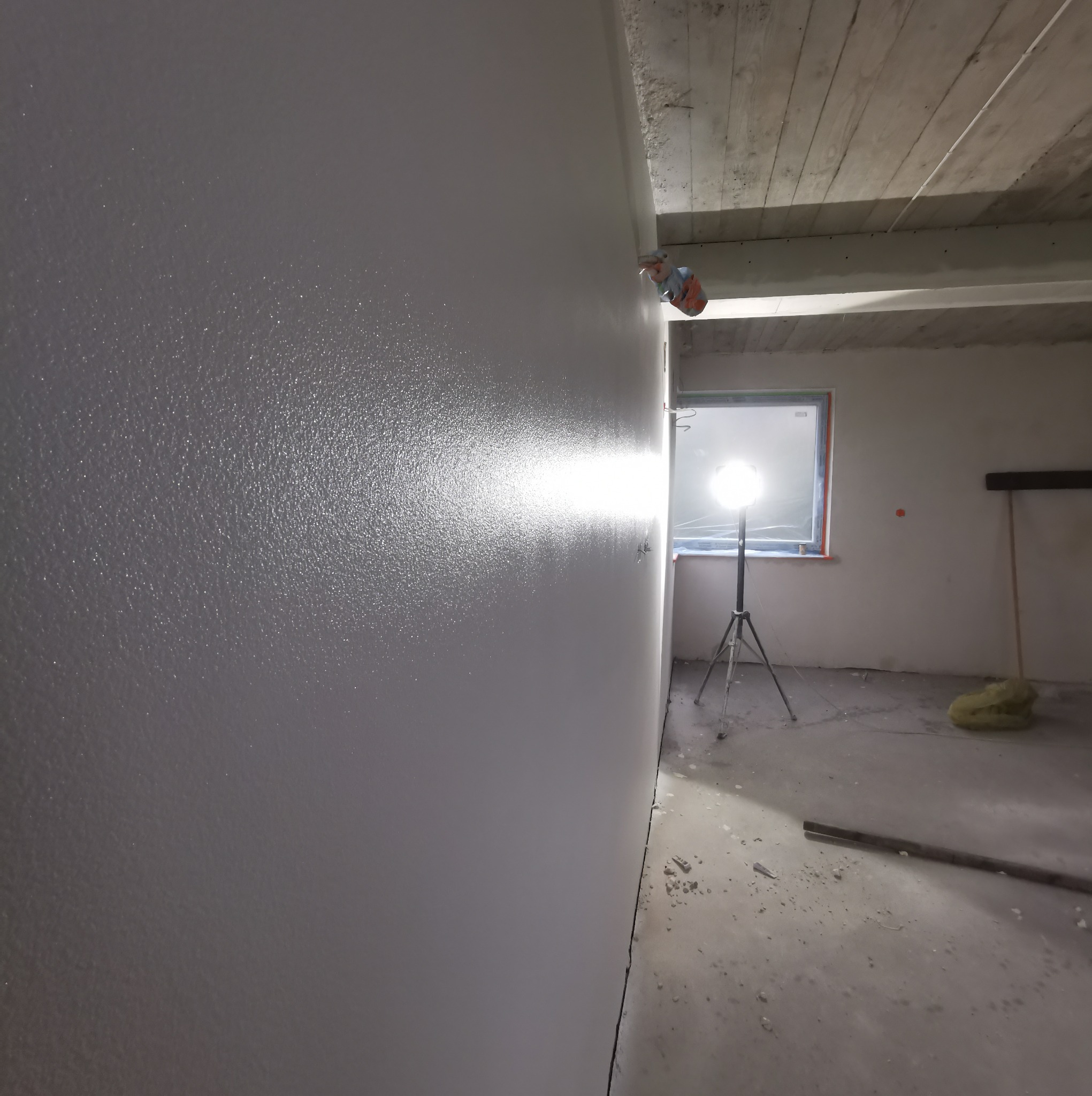 Świeżo pomalowana ściana w szarym kolorze, widoczna faktura, w tle lampa budowlana na statywie oświetlająca pomieszczenie w trakcie prac.