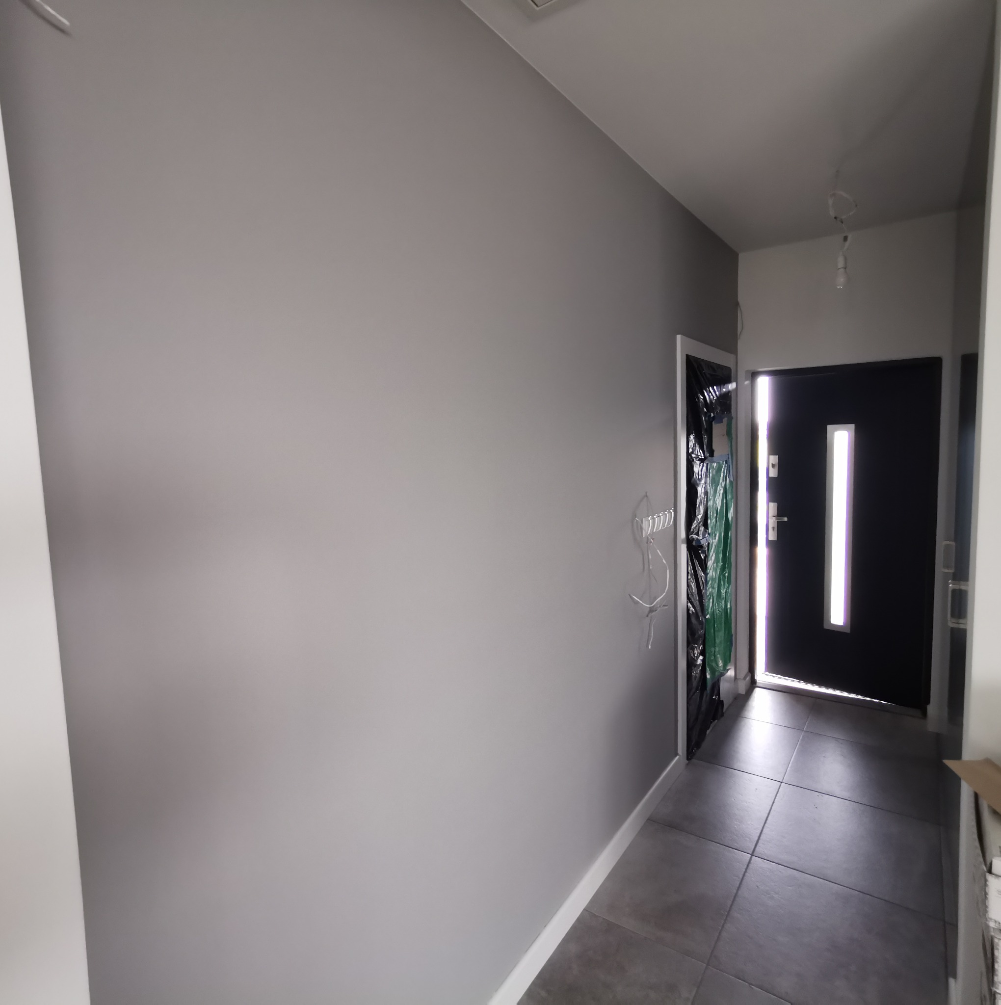 Świeżo pomalowany korytarz w szarym odcieniu, widoczna czarna okleina zabezpieczająca ościeżnicę drzwi wejściowych i szara podłoga z płytek.