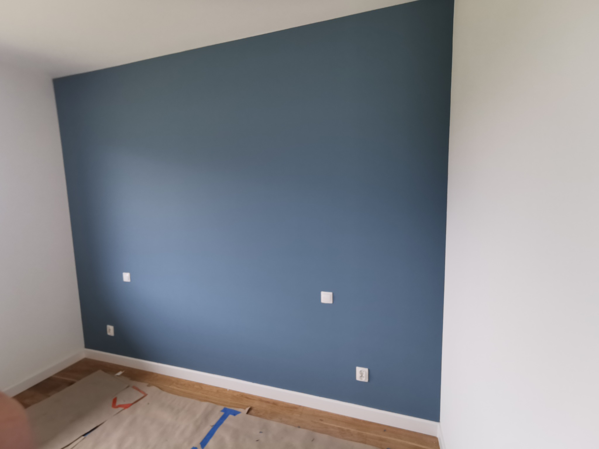 Pomieszczenie po świeżym pomalowaniu ścian na kolor granatowy, zabezpieczone podłogi kartonem i folią malarską, widoczne gniazdka elektryczne.