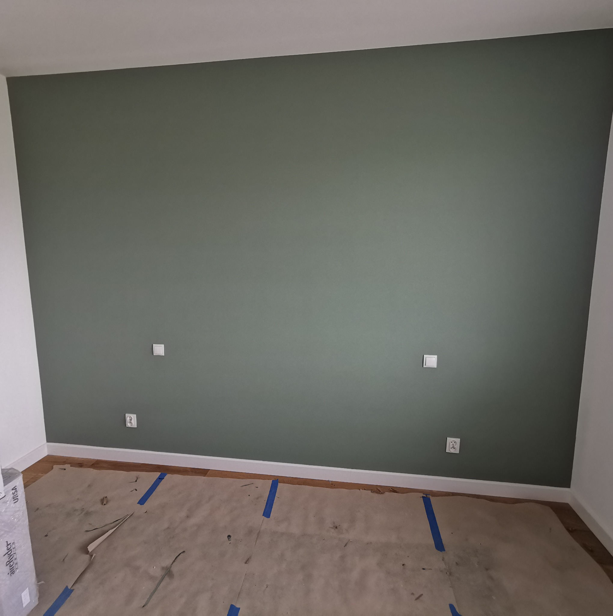 Świeżo pomalowana ściana w odcieniach zieleni z białymi listwami przypodłogowymi i zabezpieczoną podłogą, widoczne gniazdka elektryczne.