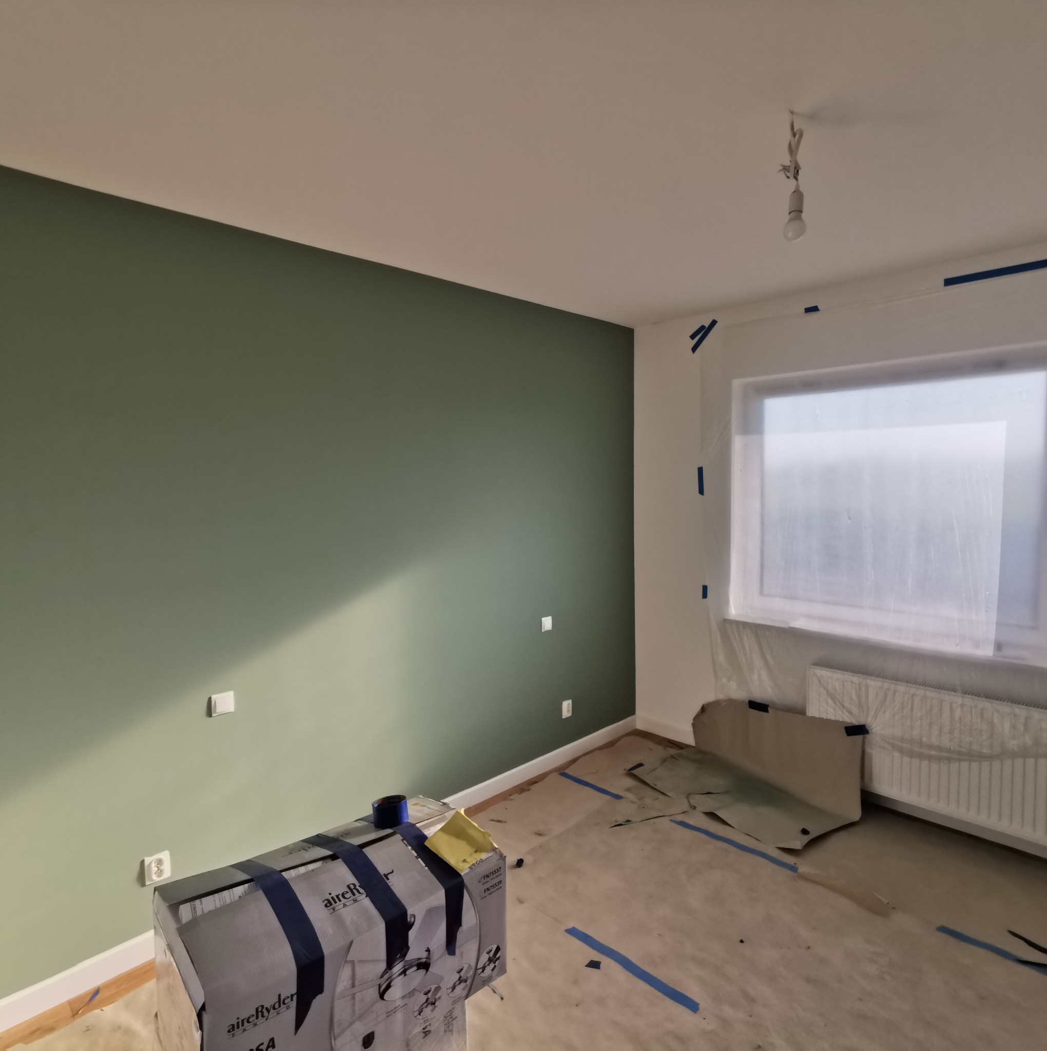 Pokój w trakcie malowania: ściana w odcieniach zieleni, zabezpieczone okno folią, zaklejona podłoga, widoczny karton z wentylatorem sufitowym.