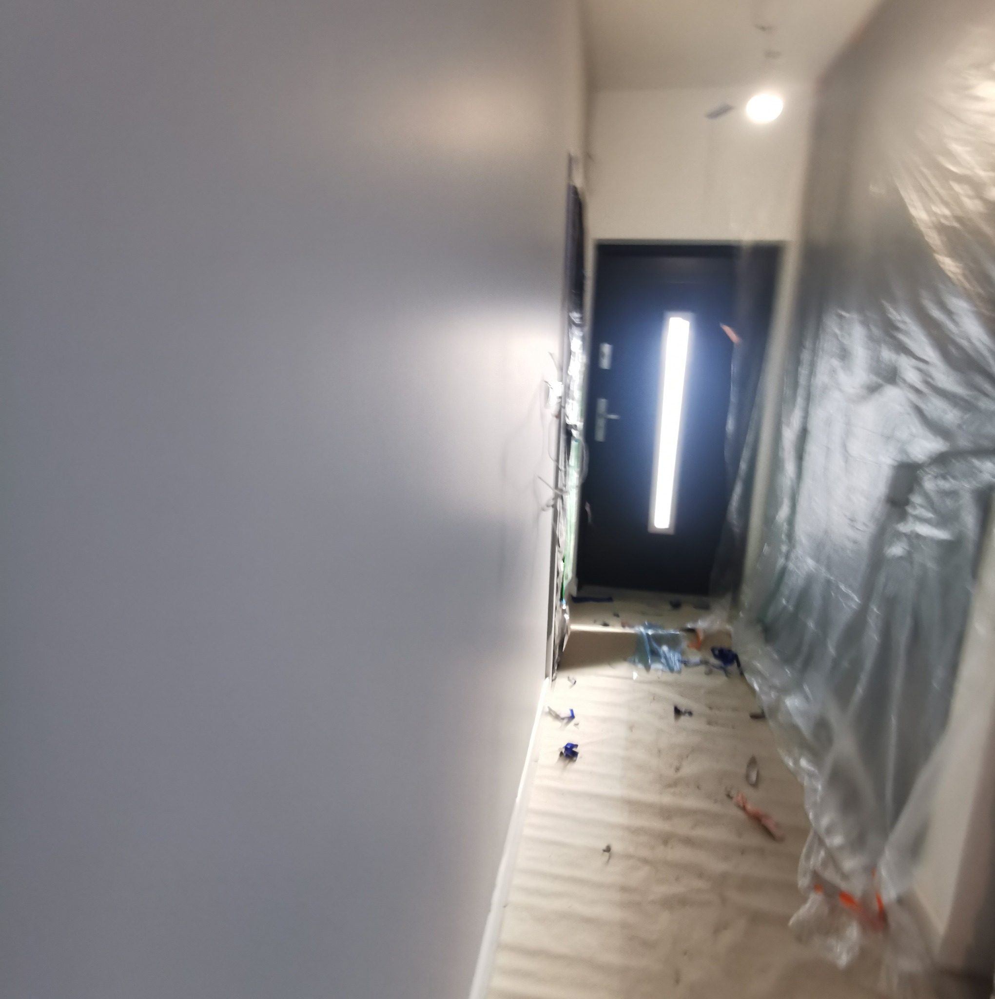 Korytarz w trakcie malowania na szaro, ściany zabezpieczone folią malarską, widoczne ślady farby na podłodze i listwy przypodłogowe.