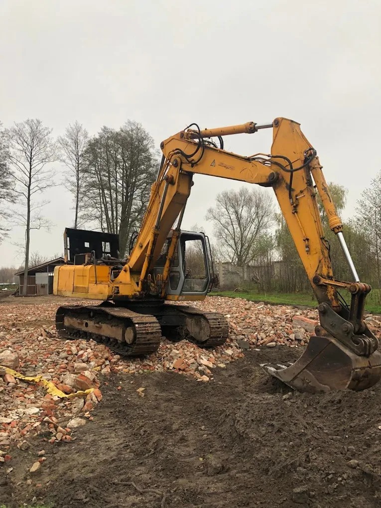 Żółta koparka gąsienicowa na placu budowy, z gruzem po rozbiórce budynku, w tle drzewa i pochmurne niebo.
