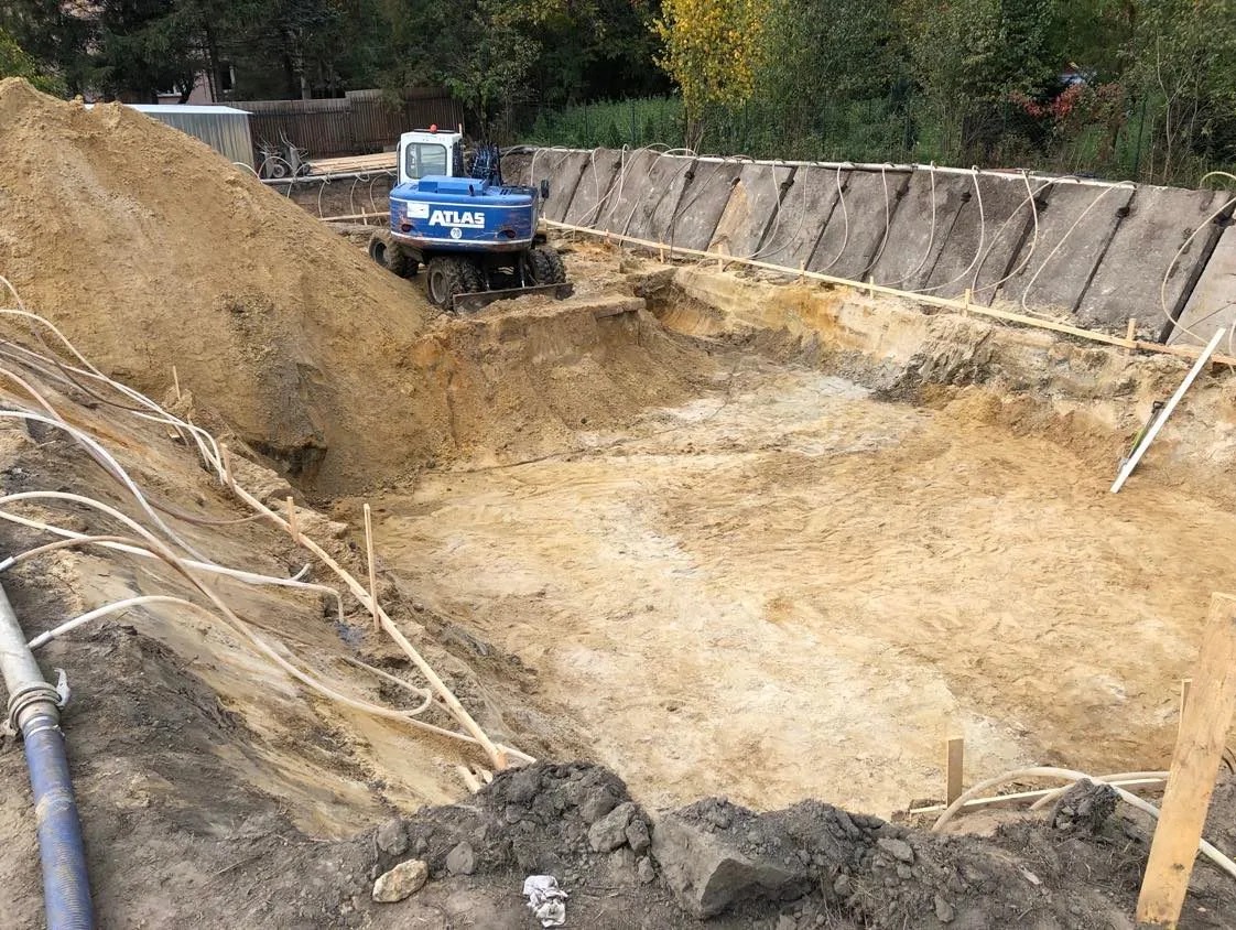 Niebieska koparka Atlas na dnie wykopu z betonowymi płytami oporowymi i wystającymi rurami, obok hałda ziemi.