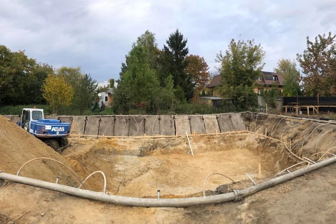 Głęboki wykop w ziemi, zabezpieczony betonowymi płytami i drewnianymi stemplami, z widocznym na skraju terenu niebieskim mini-koparką marki Atlas.