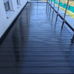 HOGAASTAR SPÓŁKA Z OGRANICZONĄ ODPOWIEDZIALNOŚCIĄ - Świeżo umyta, ciemna, kompozytowa deska tarasowa na balkonie z szklanymi barierkami, mokra od wody z widocznymi refleksami świetlnymi.