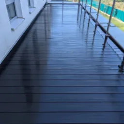 Świeżo umyta, ciemna, kompozytowa deska tarasowa na balkonie z szklanymi barierkami, mokra od wody z widocznymi refleksami świetlnymi.