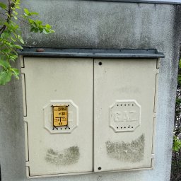 HOGAASTAR SPÓŁKA Z OGRANICZONĄ ODPOWIEDZIALNOŚCIĄ - Zabrudzona skrzynka gazowa na elewacji budynku, widoczne ślady po zaciekach i zabrudzeniach atmosferycznych, obok rosnąca zieleń.