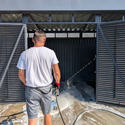 HOGAASTAR SPÓŁKA Z OGRANICZONĄ ODPOWIEDZIALNOŚCIĄ - Mężczyzna w szarych spodenkach i białej koszulce czyści podłogę w garażu za pomocą myjki ciśnieniowej, w tle otwarte, metalowe, żaluzjowe drzwi.