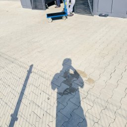 HOGAASTAR SPÓŁKA Z OGRANICZONĄ ODPOWIEDZIALNOŚCIĄ - Cień osoby na kostce brukowej w słoneczny dzień, w tle fragment wózka transportowego i metalowej konstrukcji.