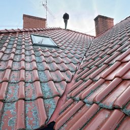 HOGAASTAR SPÓŁKA Z OGRANICZONĄ ODPOWIEDZIALNOŚCIĄ - Czerwona dachówka z widocznym mchem i człowiekiem na szczycie, komin oraz antena w tle. Dachówka po prawej stronie wygląda na świeżo umytą.
