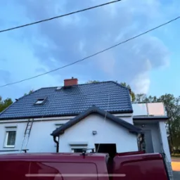 Dom z ciemnym dachem po usłudze dekarskiej. Na ścianie oparta drabina. Widoczny fragment czerwonego samochodu. Pochmurne niebo.