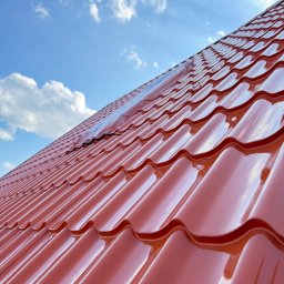 HOGAASTAR SPÓŁKA Z OGRANICZONĄ ODPOWIEDZIALNOŚCIĄ - Błyszczący, nowy dach z blachy trapezowej w odcieniach czerwieni pod błękitnym niebem z chmurami. Widoczny fragment folii ochronnej. Ujęcie z dołu.