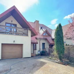 Czyszczenie dachu domu jednorodzinnego z czerwoną dachówką przez pracownika w uprzęży, za pomocą myjki ciśnieniowej. Widoczny budynek z garażem i kostka brukowa.