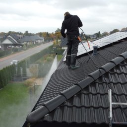 HOGAASTAR SPÓŁKA Z OGRANICZONĄ ODPOWIEDZIALNOŚCIĄ - Pracownik w czarnym kombinezonie czyści dach z czarnej dachówki, stojąc na nim. W tle widoczne domy i zieleń. Na dachu panele słoneczne i drabina.