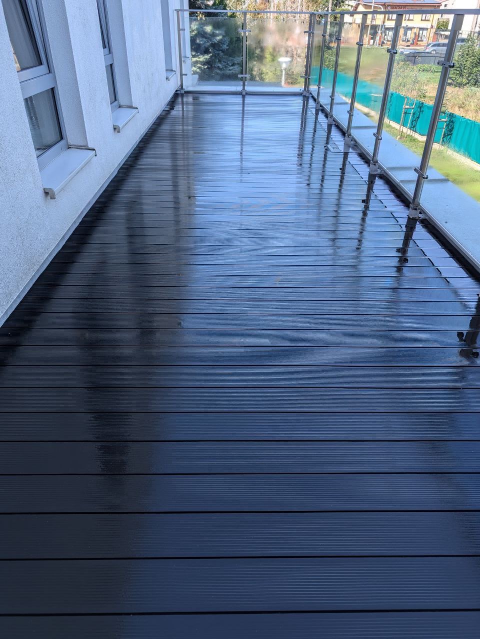 Świeżo umyta, ciemna, kompozytowa deska tarasowa na balkonie z szklanymi barierkami, mokra od wody z widocznymi refleksami świetlnymi.