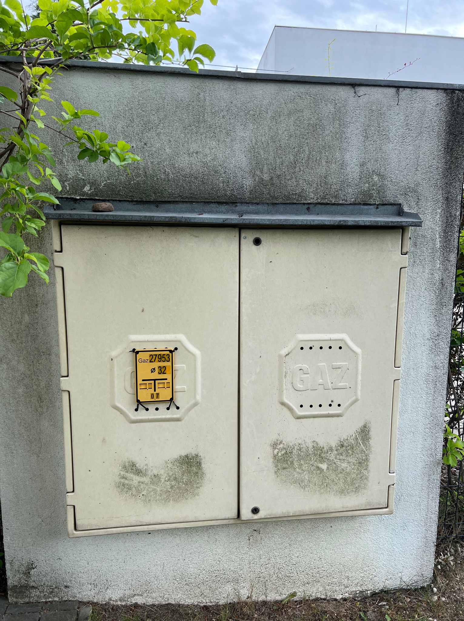 Zabrudzona skrzynka gazowa na elewacji budynku, widoczne ślady po zaciekach i zabrudzeniach atmosferycznych, obok rosnąca zieleń.