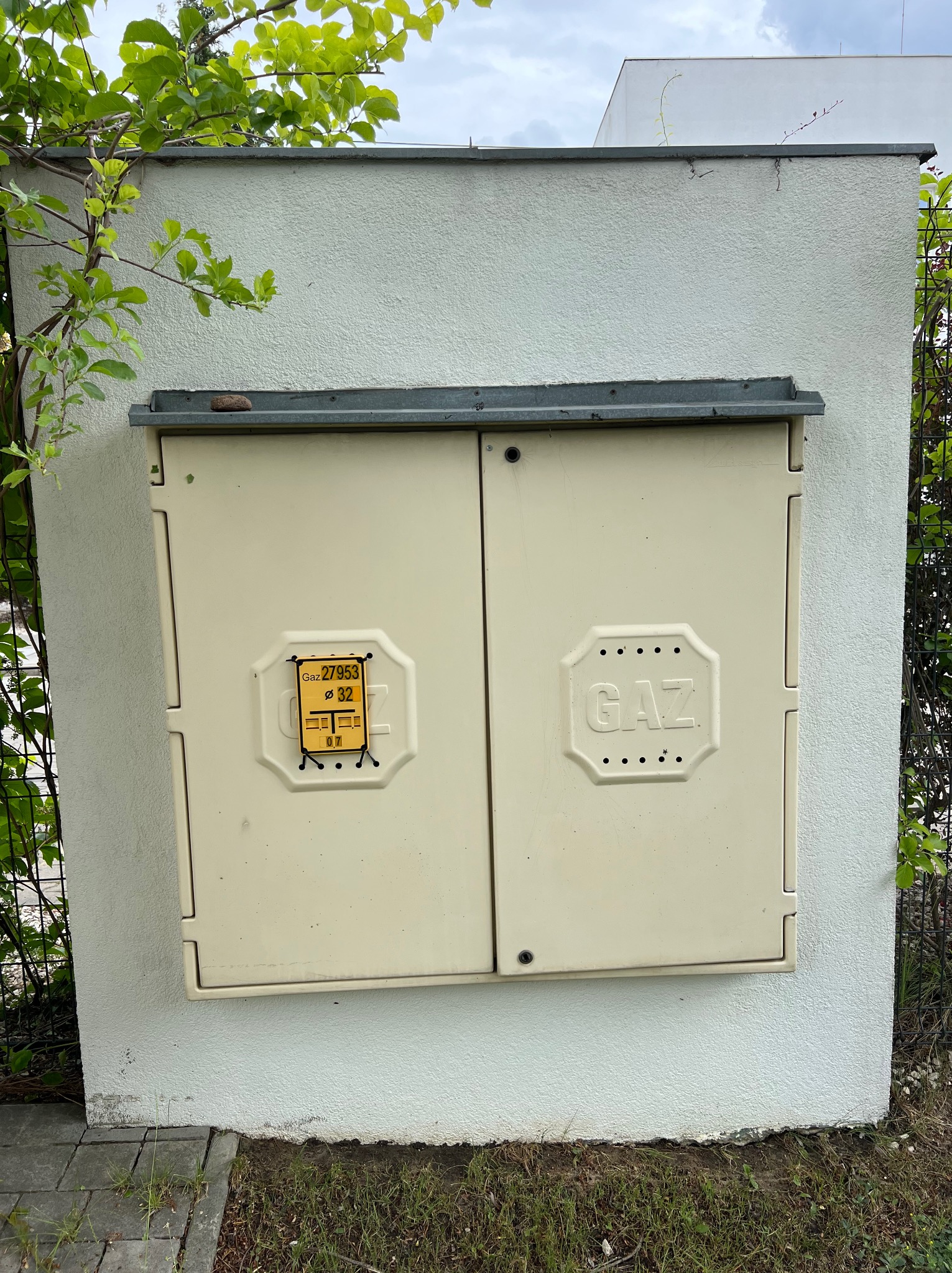 Skrzynka gazowa na elewacji budynku, z żółtą naklejką informacyjną i roślinnością w tle.