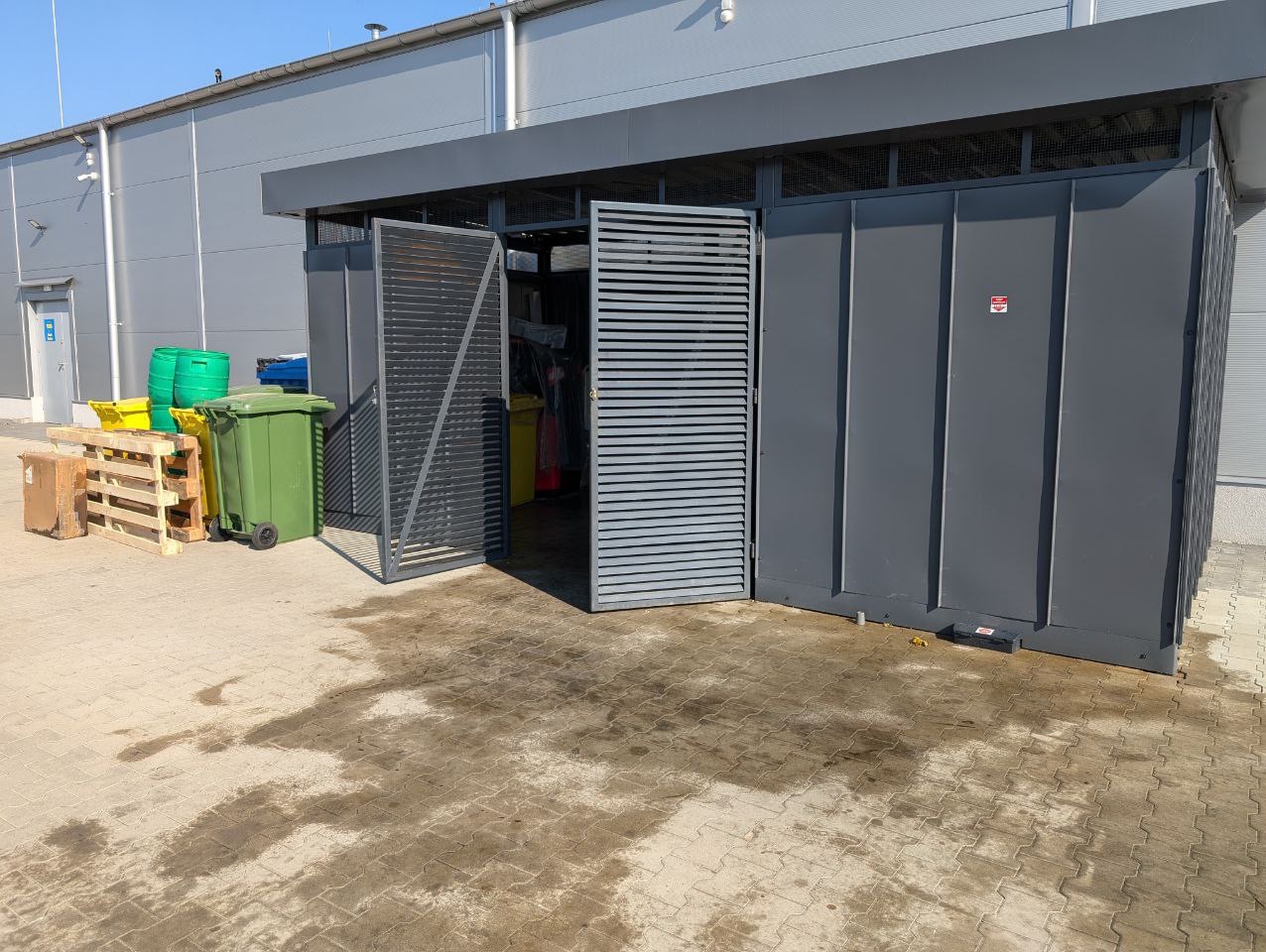 Zewnętrzna wiata śmietnikowa z metalowymi żaluzjowymi drzwiami, obok ustawione kontenery na śmieci i palety, na kostce brukowej widoczne ślady po wodzie.