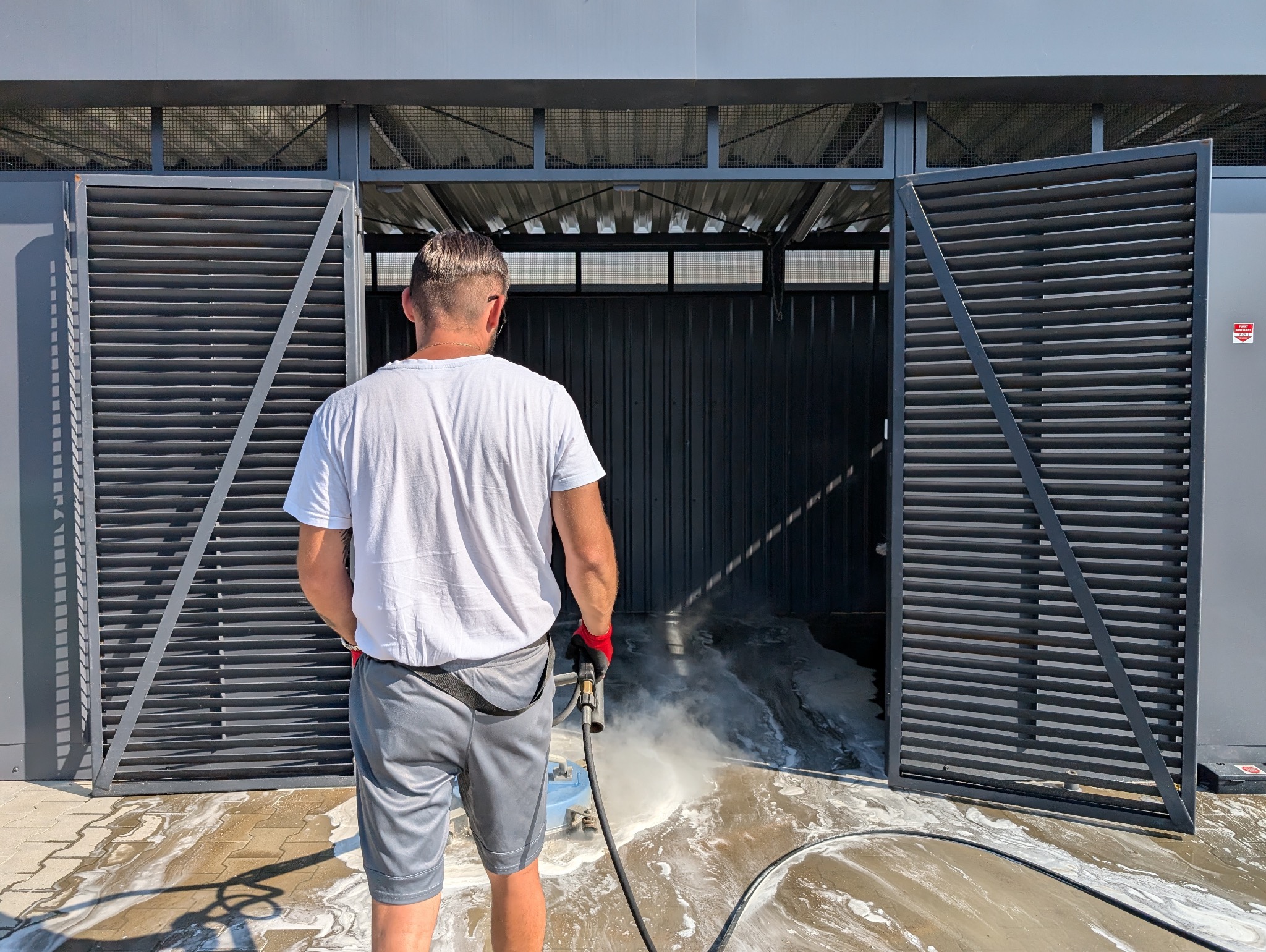 Mężczyzna w szarych spodenkach i białej koszulce czyści podłogę w garażu za pomocą myjki ciśnieniowej, w tle otwarte, metalowe, żaluzjowe drzwi.