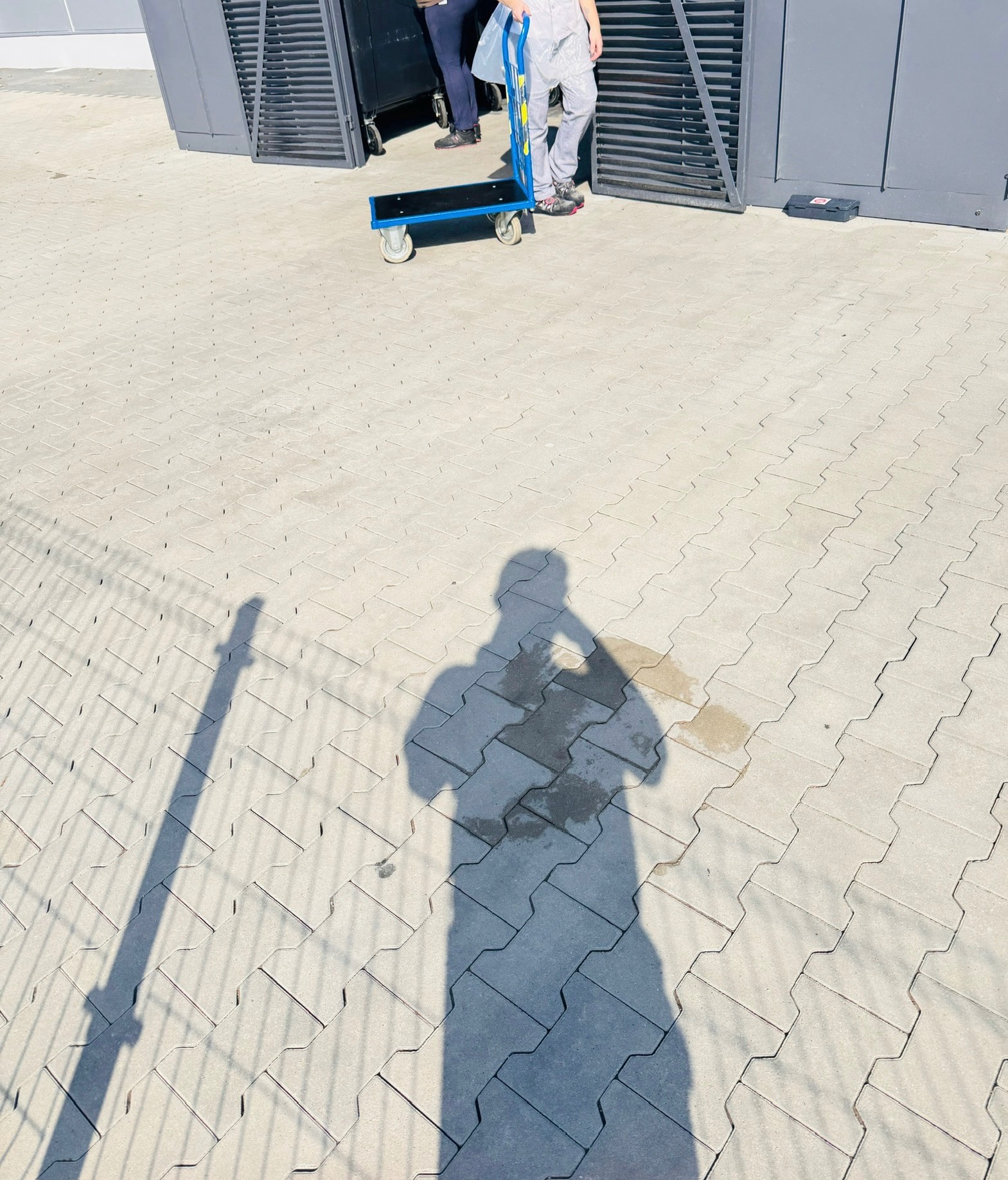 Cień osoby na kostce brukowej w słoneczny dzień, w tle fragment wózka transportowego i metalowej konstrukcji.