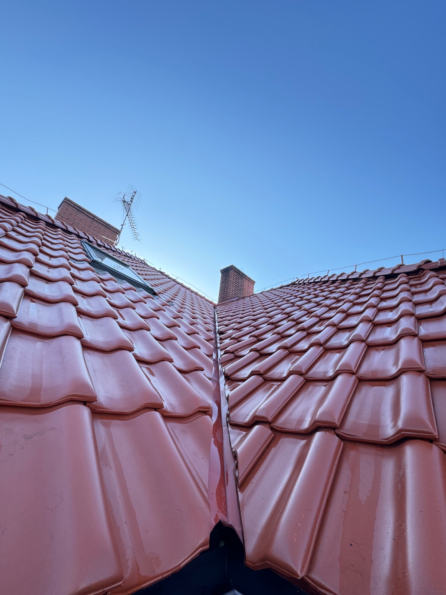 Czerwona, błyszcząca dachówka ceramiczna po deszczu, widok z dołu na dach z kominem, oknem dachowym i anteną na tle błękitnego nieba.
