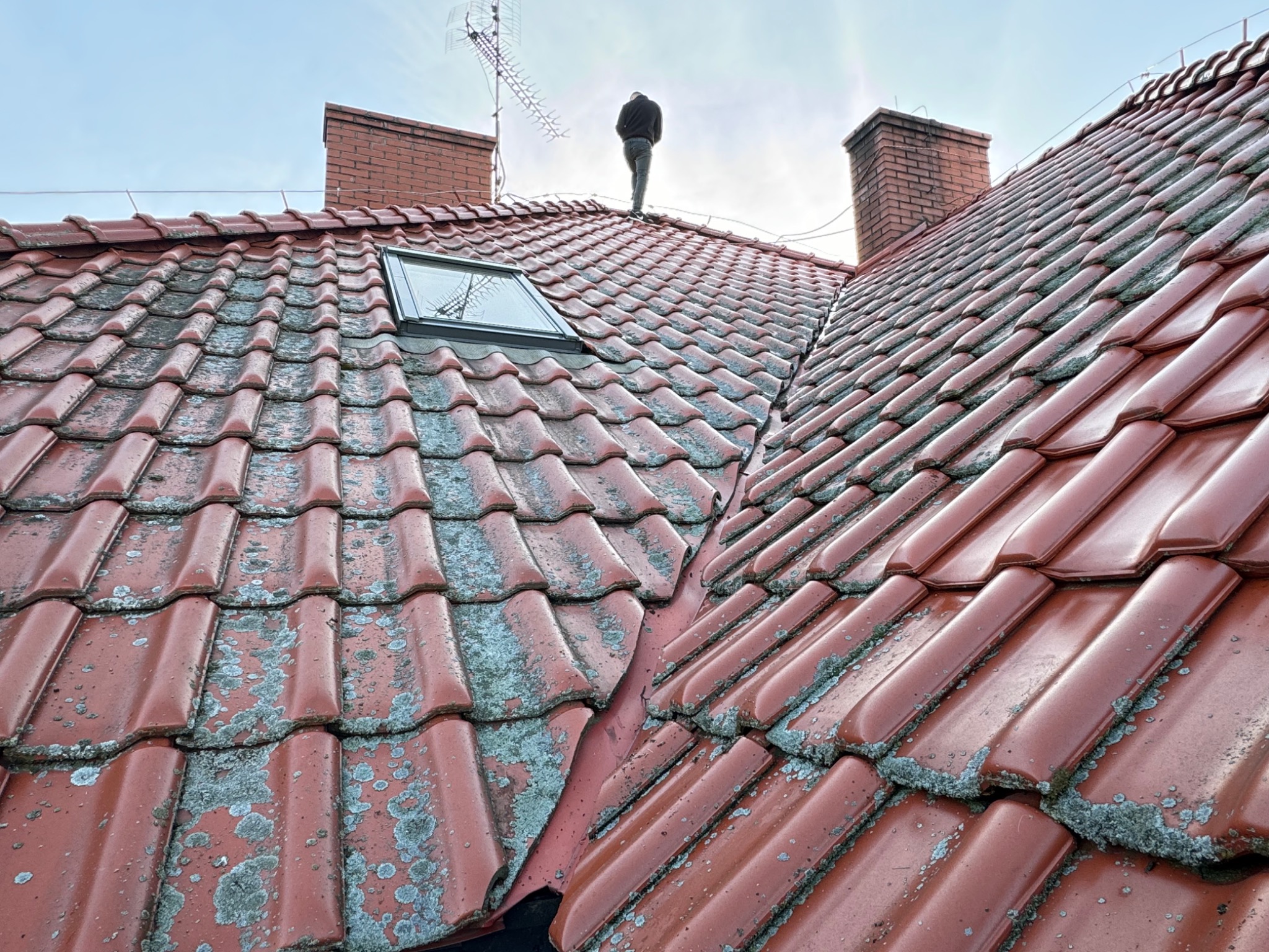 Czerwona dachówka z widocznym mchem i człowiekiem na szczycie, komin oraz antena w tle. Dachówka po prawej stronie wygląda na świeżo umytą.