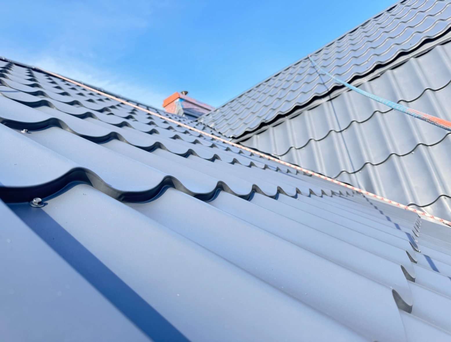 Szary dach z blachy trapezowej pod błękitnym niebem, widoczny komin zabezpieczony folią. Ujęcie z dołu eksponuje regularny wzór pokrycia dachowego.