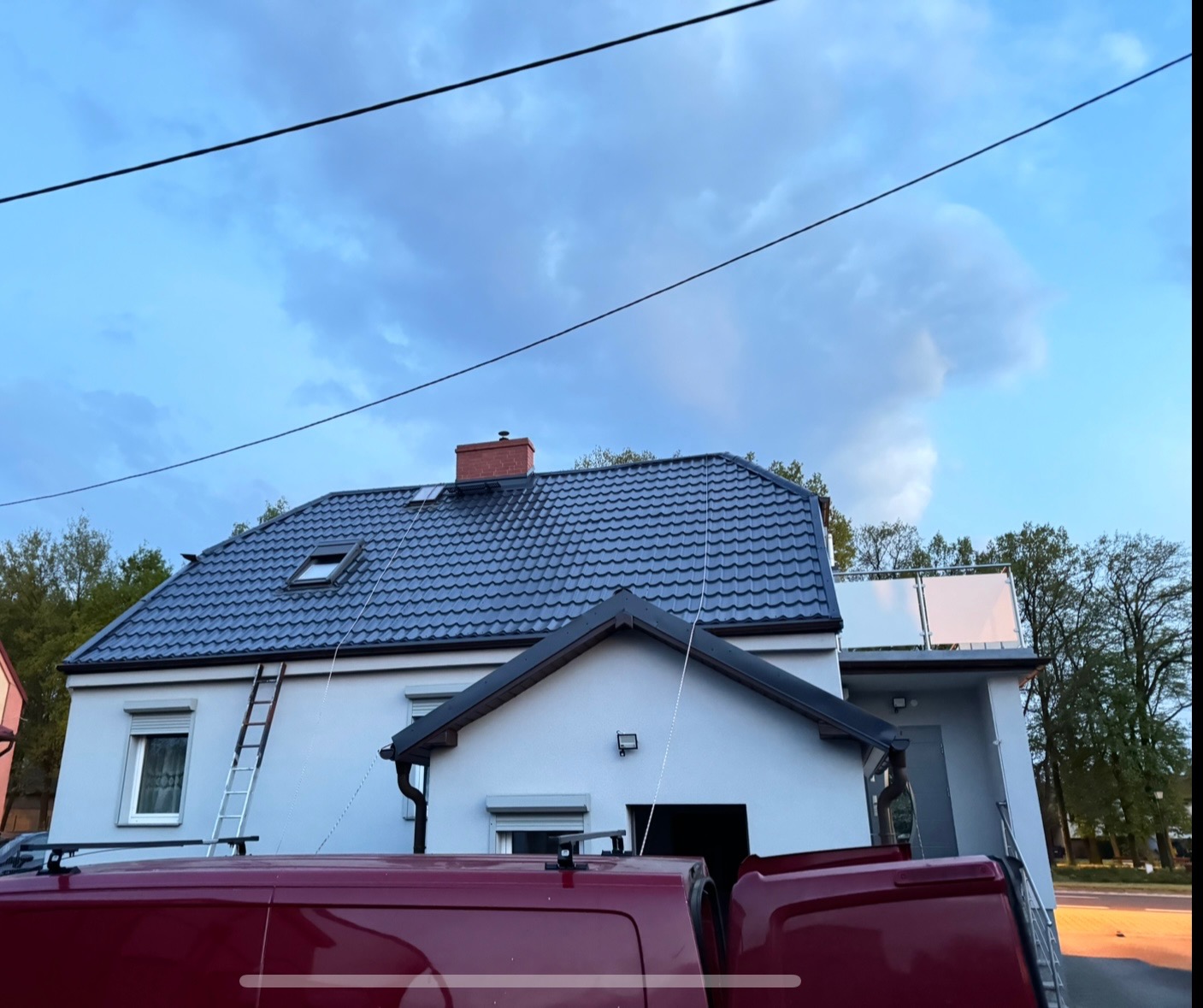 Dom z ciemnym dachem po usłudze dekarskiej. Na ścianie oparta drabina. Widoczny fragment czerwonego samochodu. Pochmurne niebo.