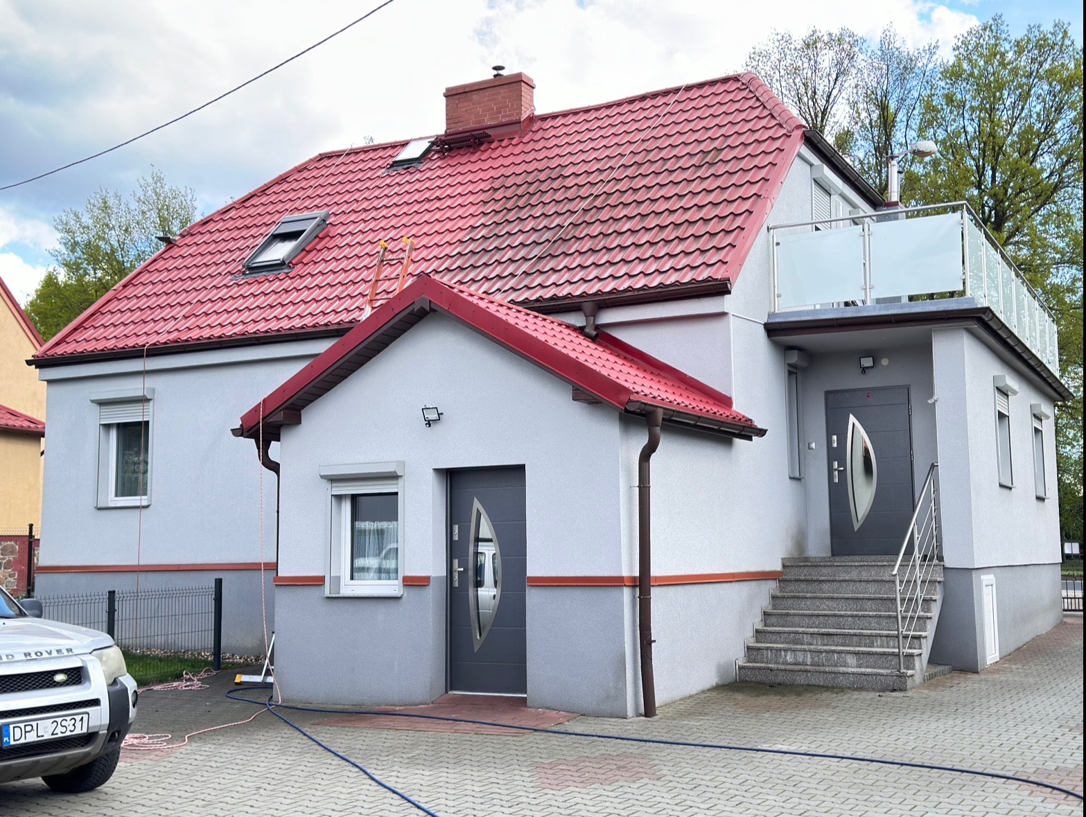 Dom jednorodzinny z czerwonym dachem po czyszczeniu, widoczne ślady wody na dachu, drabina oparta o dach, szary samochód na pierwszym planie, kostka brukowa.