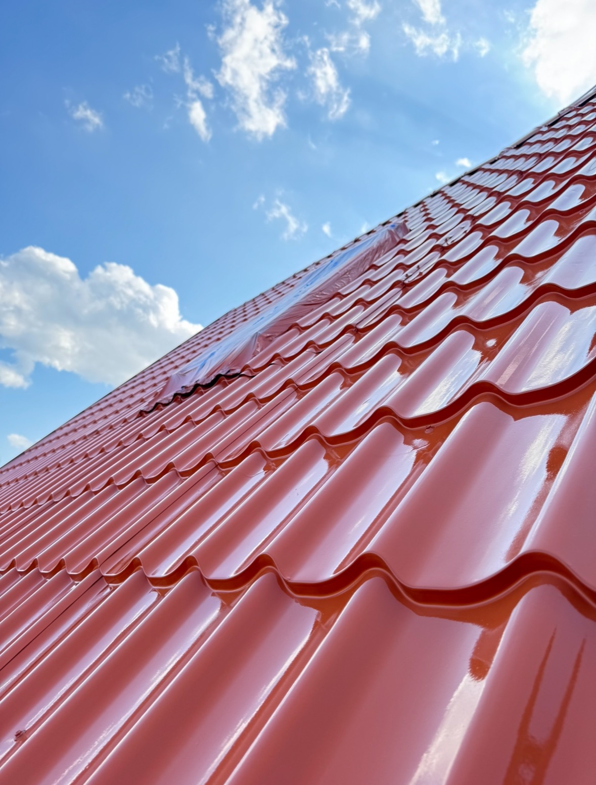 Błyszczący, nowy dach z blachy trapezowej w odcieniach czerwieni pod błękitnym niebem z chmurami. Widoczny fragment folii ochronnej. Ujęcie z dołu.