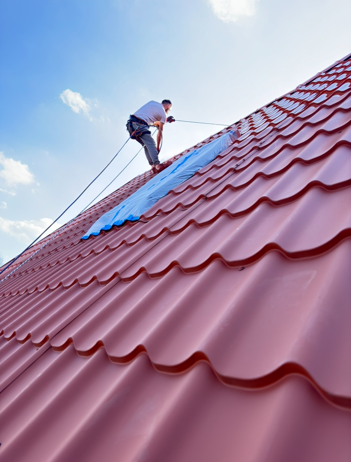 Pracownik zabezpieczony linami na dachu z blachodachówki w trakcie pracy, fragment dachu przykryty folią. Ujęcie z dołu, błękitne niebo w tle.