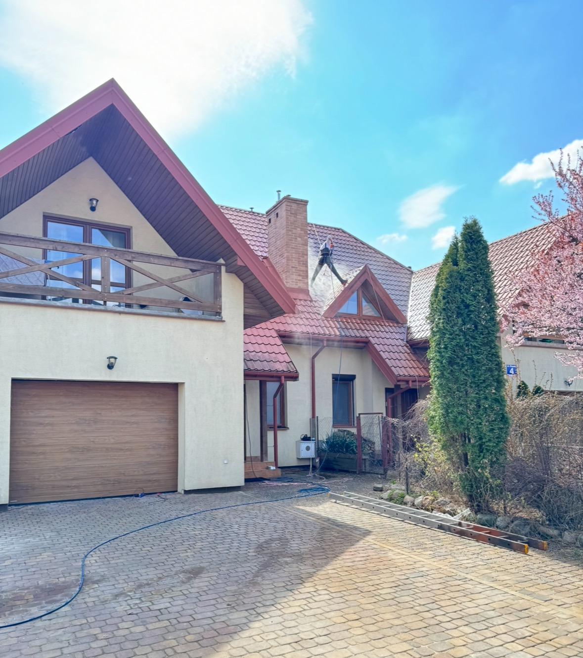 Czyszczenie dachu domu jednorodzinnego z czerwoną dachówką przez pracownika w uprzęży, za pomocą myjki ciśnieniowej. Widoczny budynek z garażem i kostka brukowa.