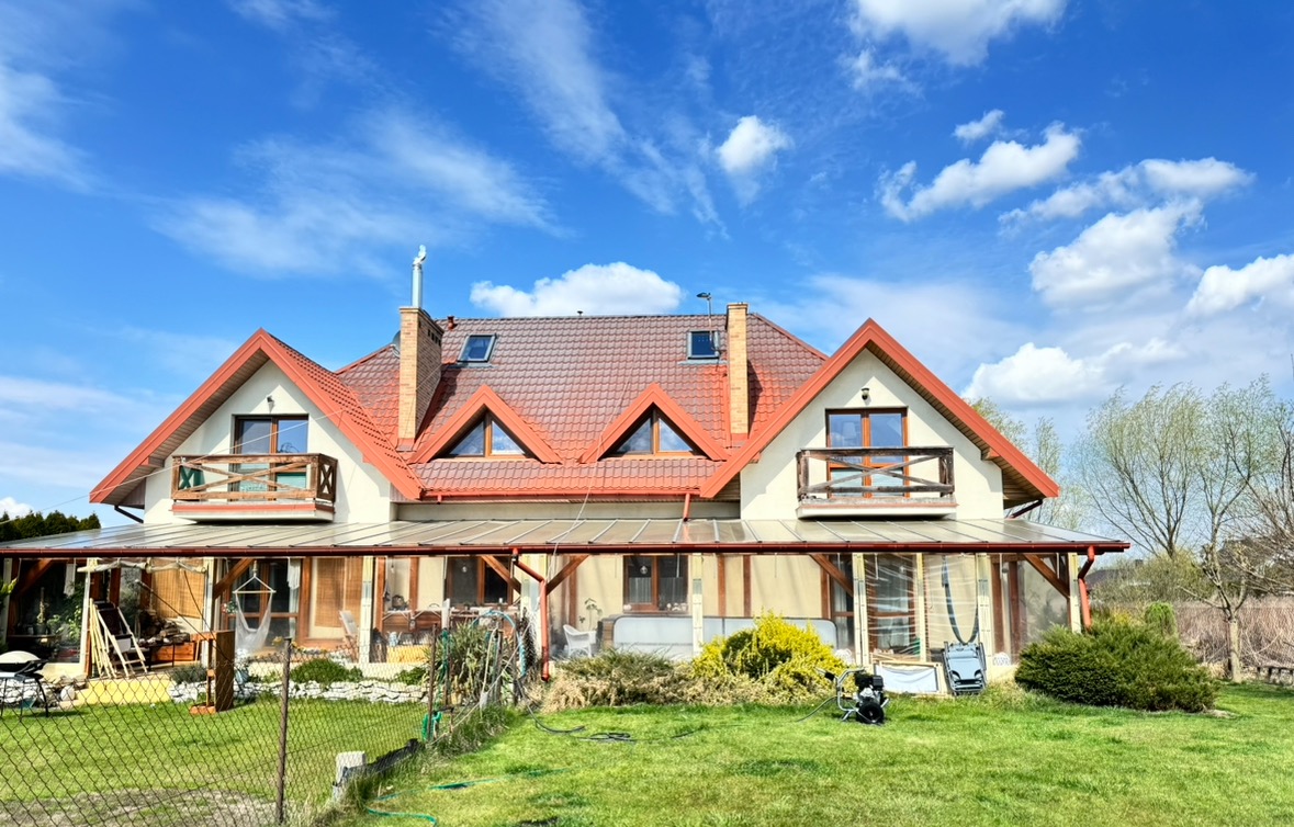 Duży dom z czerwoną dachówką i oranżerią, widziany z ogrodu w słoneczny dzień. Na trawniku stoi myjka ciśnieniowa.