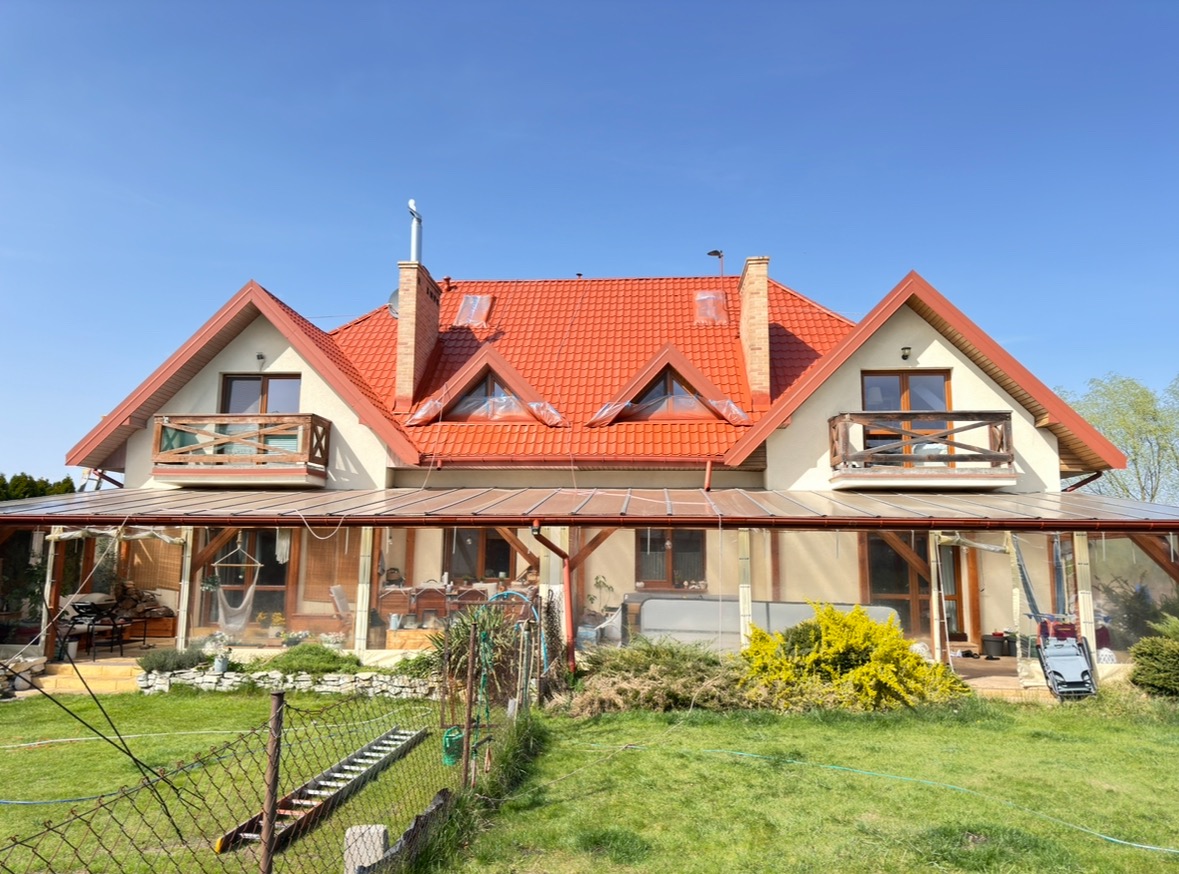 Dom jednorodzinny z czerwonym dachem przed pracami renowacyjnymi. Okna dachowe i niektóre elementy zabezpieczone folią malarską. Widoczny trawnik i ogrodzenie.