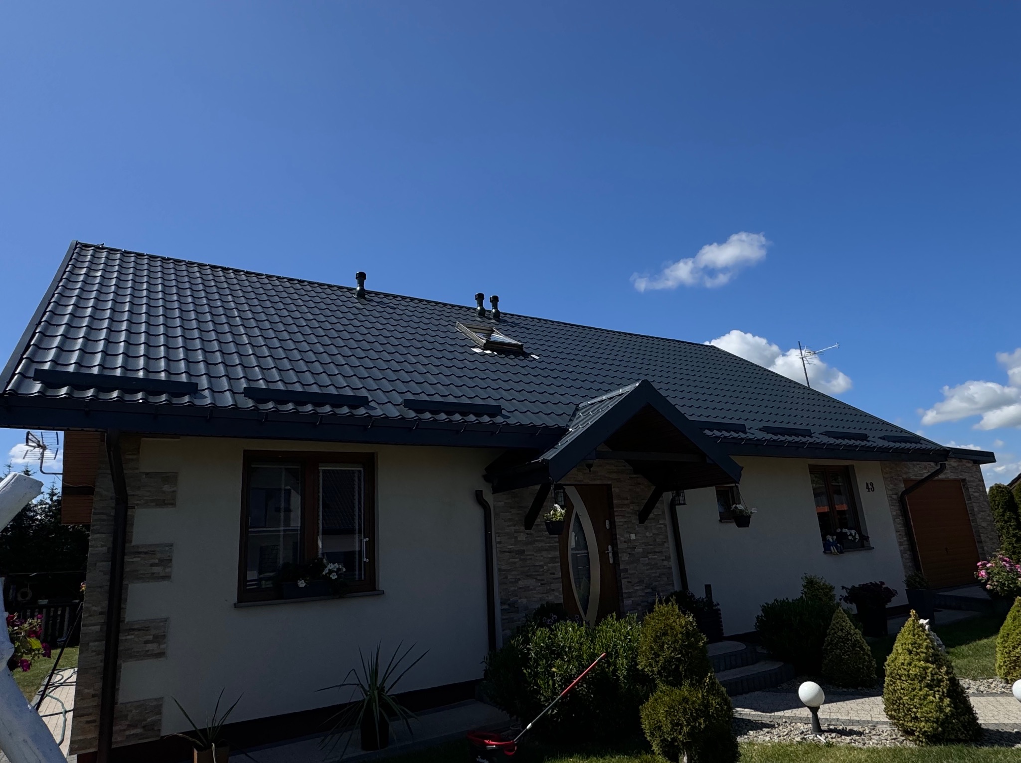Dom jednorodzinny z ciemnym, nowym dachem z dachówki ceramicznej. Widoczne okna z kwiatami i zadbany ogród. Błękitne niebo z delikatnymi obłokami w tle.