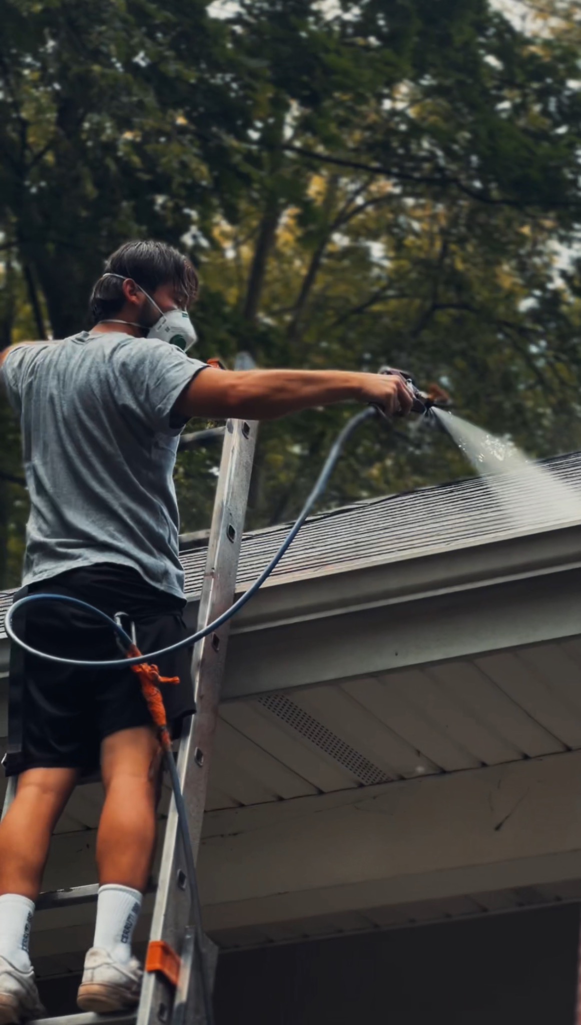 Pracownik w masce, na drabinie, czyści dach strumieniem wody pod ciśnieniem. Widoczne drzewa w tle. Ujęcie z dołu, podkreślające wysokość pracy.