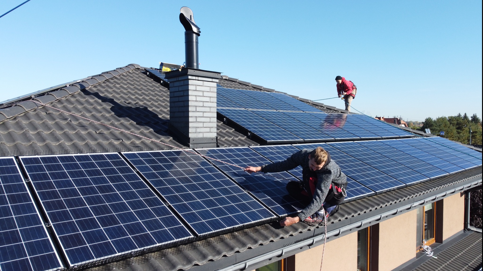 Montaż paneli słonecznych na dachu domu jednorodzinnego. Dwóch monterów zabezpieczonych linami. Dach pokryty ciemną dachówką ceramiczną, komin z cegły.
