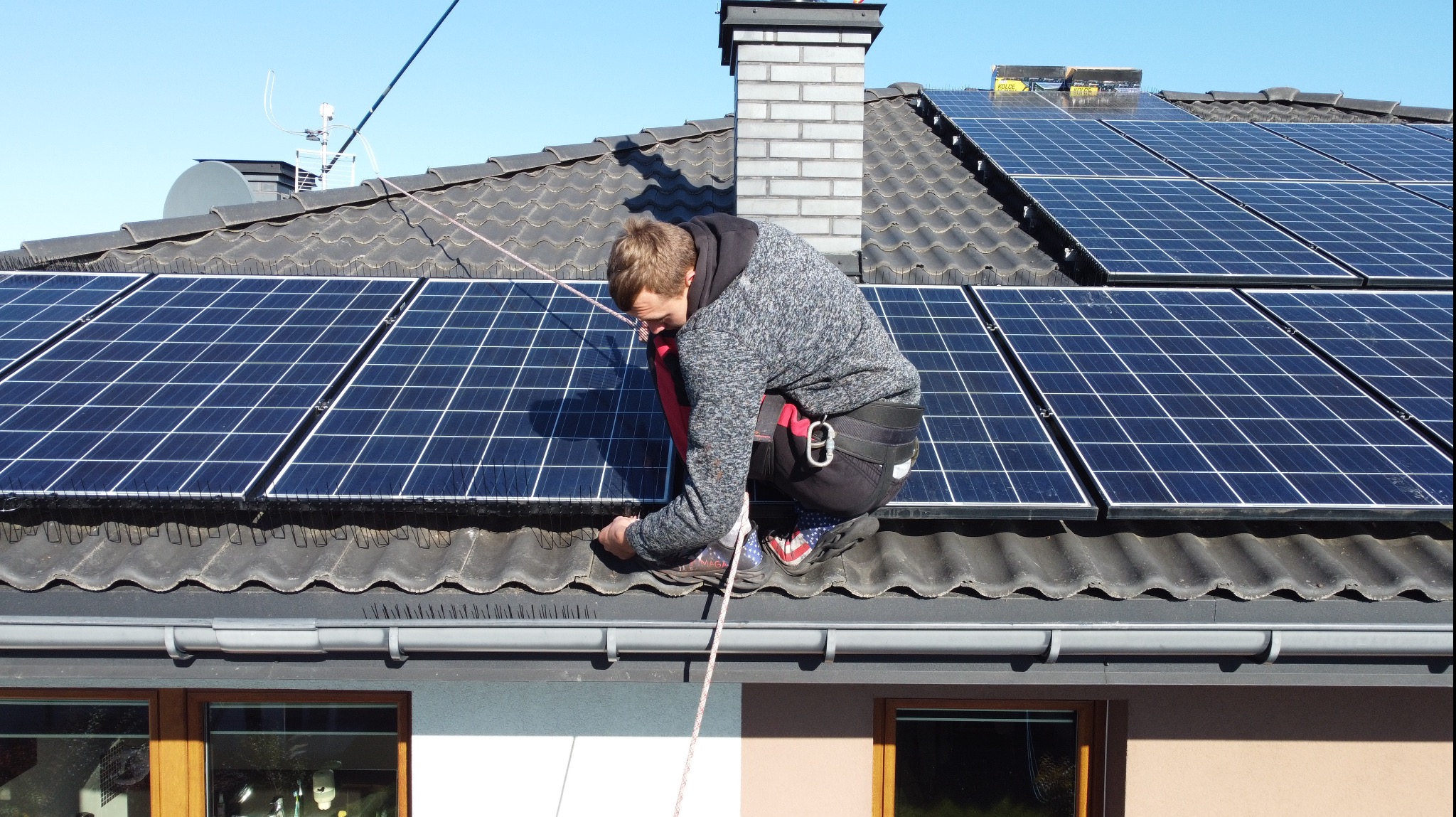Instalator w uprzęży montuje panele słoneczne na ciemnym, spadzistym dachu z ceramiczną dachówką. Widoczne zabezpieczenia przeciw ptakom i komin. Błękitne niebo w tle.