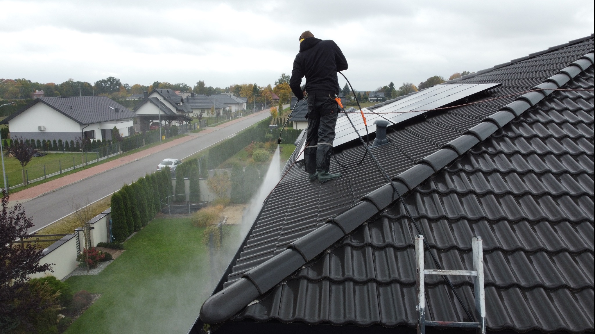 Pracownik w czarnym kombinezonie czyści dach z czarnej dachówki, stojąc na nim. W tle widoczne domy i zieleń. Na dachu panele słoneczne i drabina.