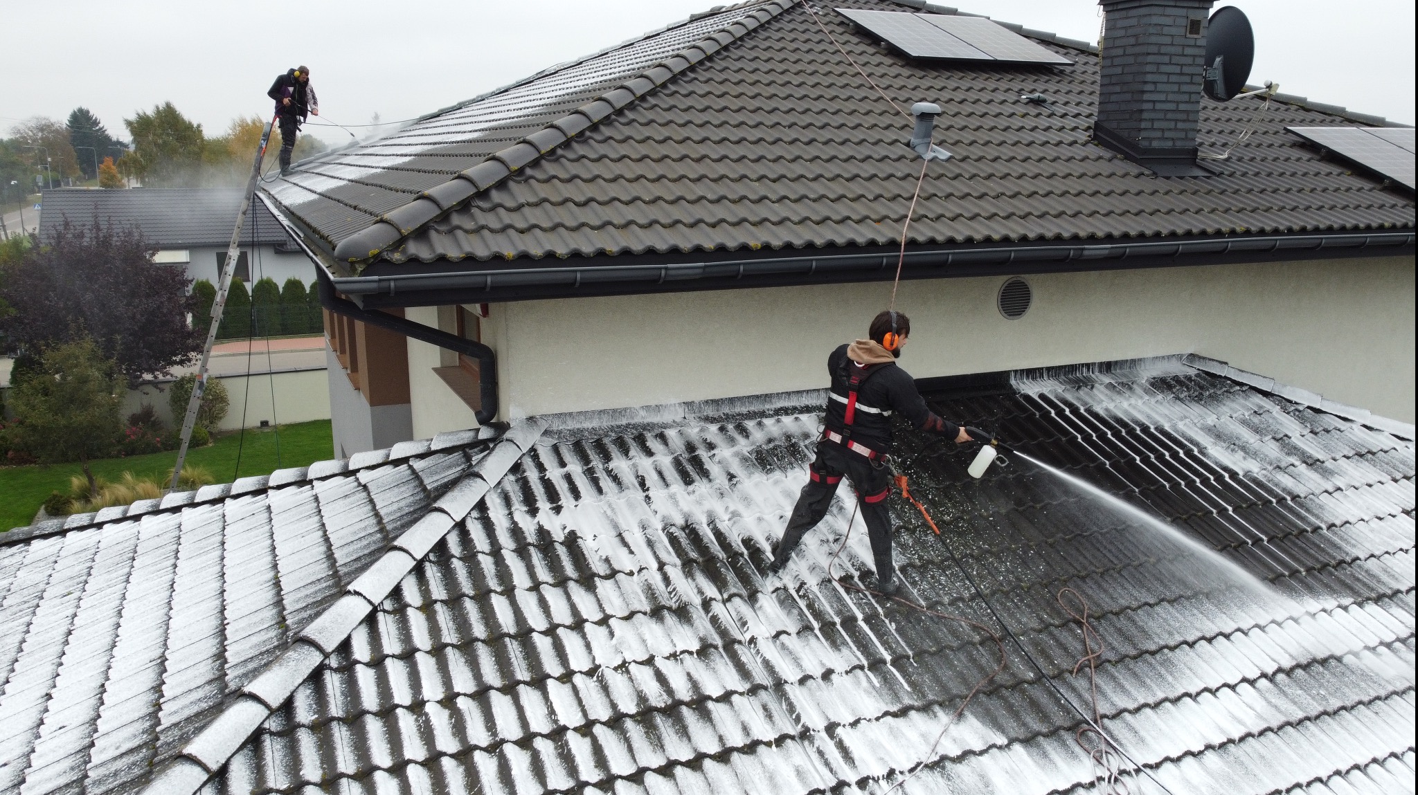 Dwóch pracowników w uprzężach czyści dach z ciemnej dachówki za pomocą myjek ciśnieniowych. Widoczne panele słoneczne i drabina oparta o budynek.