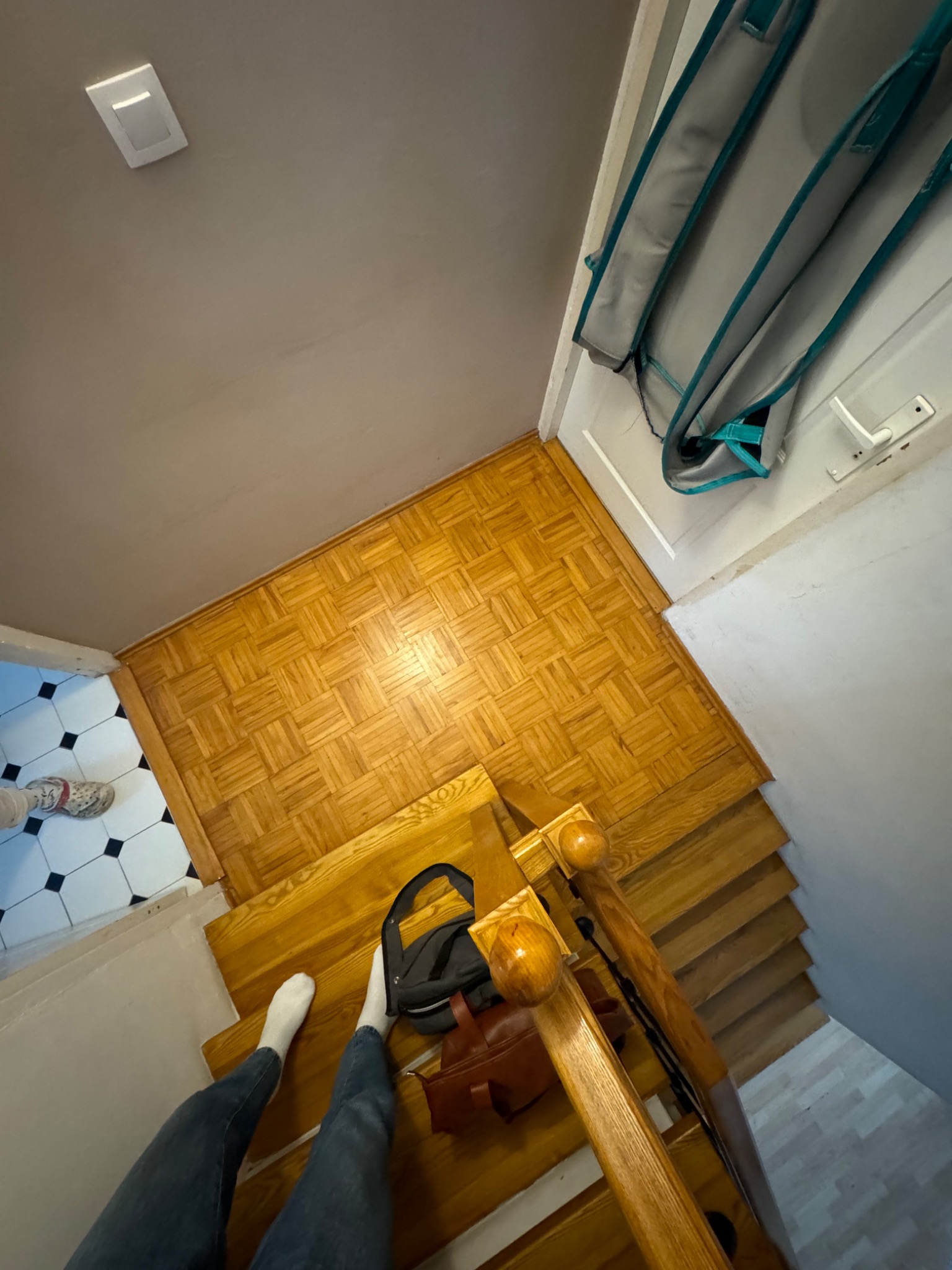 Widok z góry na drewniane schody z poręczą, fragment parkietu i kafelków. Na schodach leżą torby, a na pierwszym planie widać nogi osoby w jeansach i białych skarpetkach.