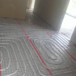 Białe rury systemu ogrzewania podłogowego ułożone na siatce w trakcie montażu, widoczne czerwone pasy taśmy mocującej, surowy stan wykończenia wnętrza.