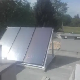 Kolektory słoneczne zamontowane na płaskim dachu budynku, widoczne elementy instalacji i komin wentylacyjny, w tle zieleń drzew i budynki.