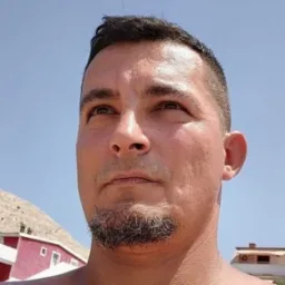 Portret mężczyzny z brodą spoglądającego w górę, na tle błękitnego nieba i częściowo widocznego budynku z czerwonymi ścianami. Ujęcie z dołu.