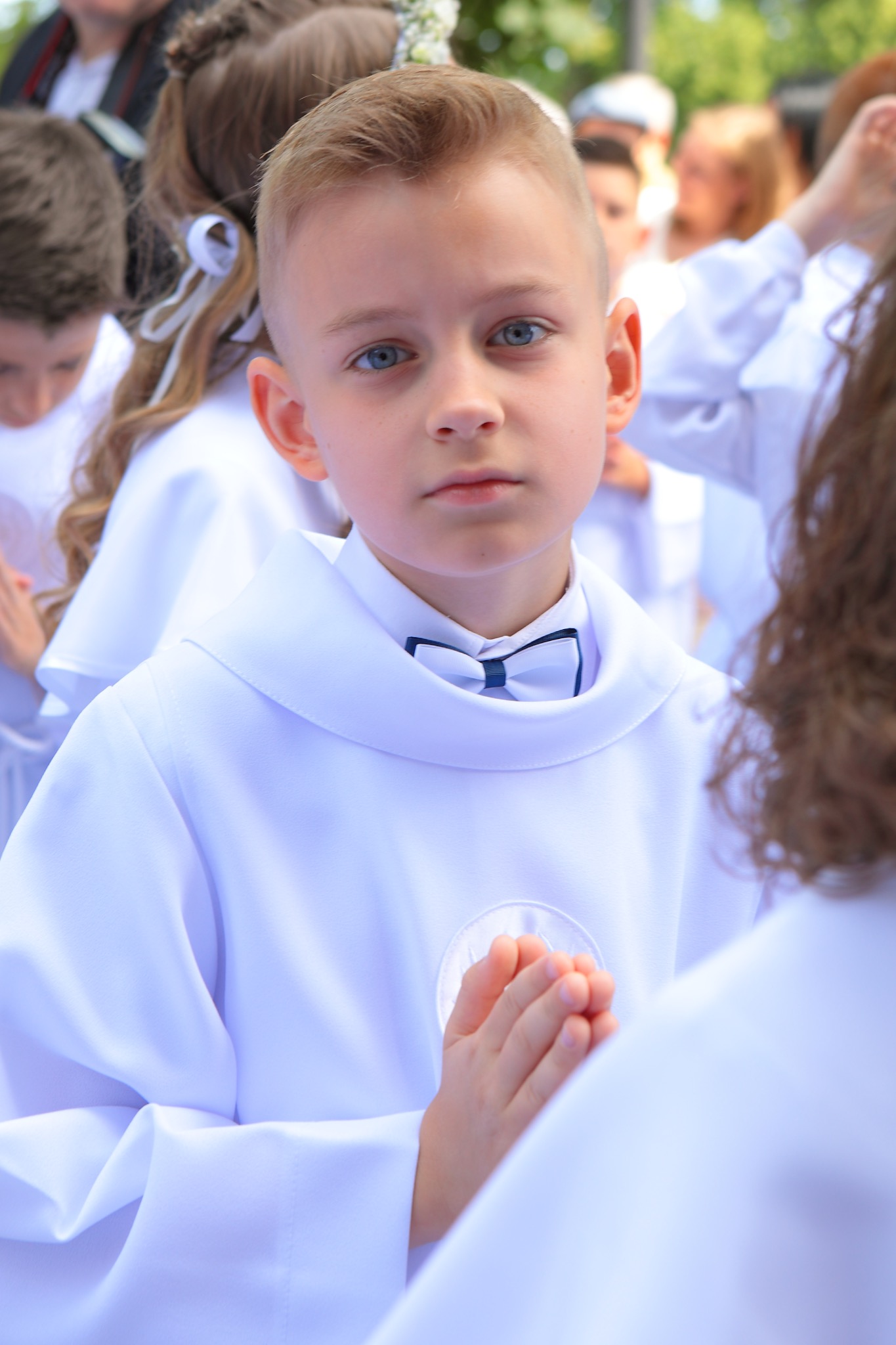 Chłopiec w białej albie komunijnej i z granatową muchą, z rękami złożonymi do modlitwy, na tle grupy dzieci w podobnych strojach.