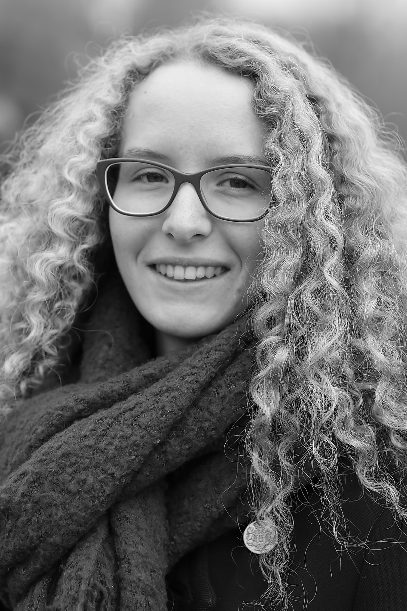 Portret uśmiechniętej młodej kobiety w okularach i szaliku, z burzą kręconych włosów, w czerni i bieli.