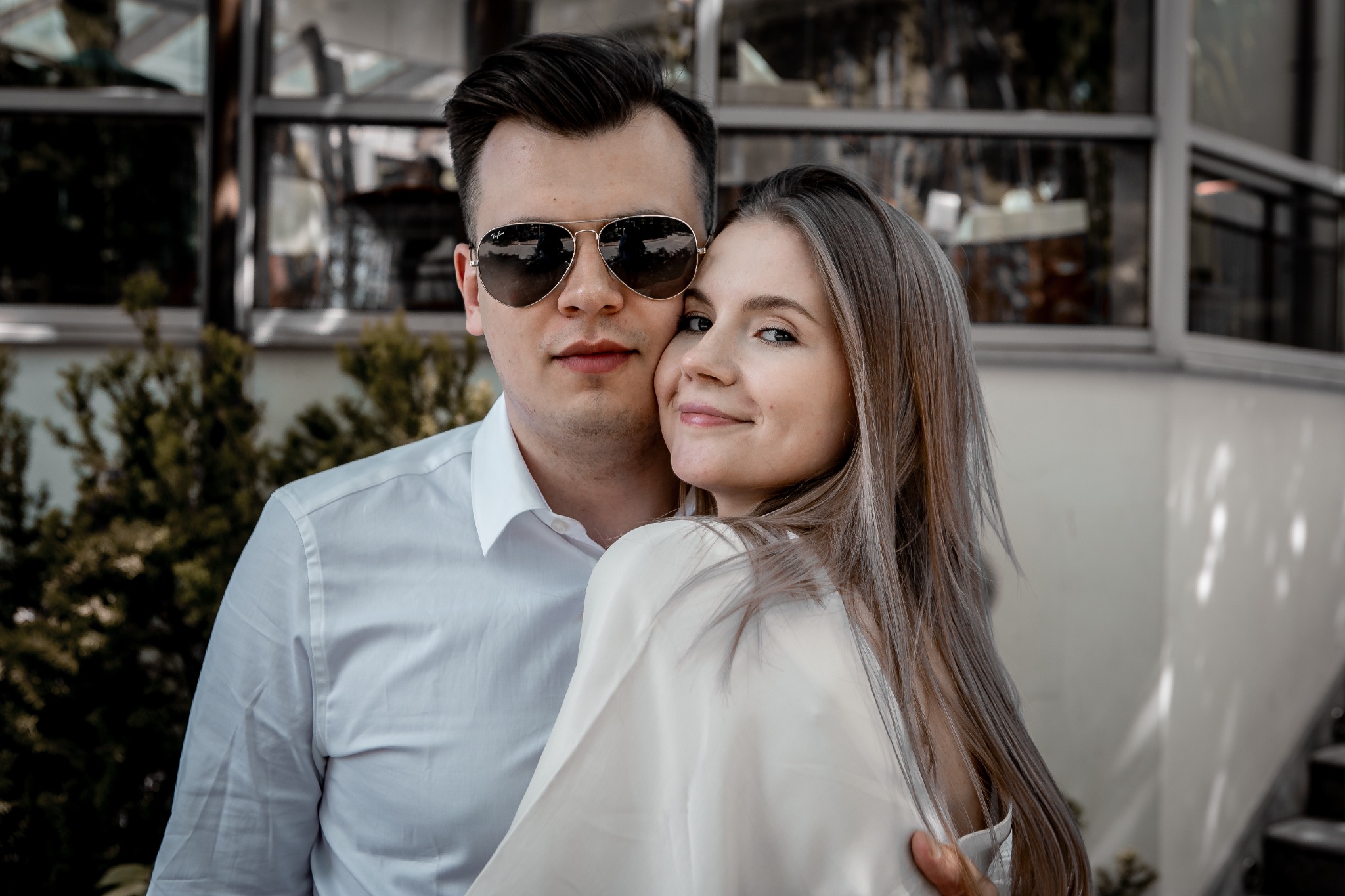 Uśmiechnięta para w objęciach na tle budynku z dużymi oknami. Mężczyzna w okularach przeciwsłonecznych, kobieta w jasnej bluzce. Romantyczna sesja w plenerze.